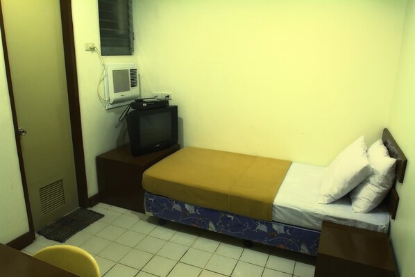 Una habitación pequeña con una cama individual, un televisor sobre un soporte de madera, una ventana con aire acondicionado y suelo de baldosas.