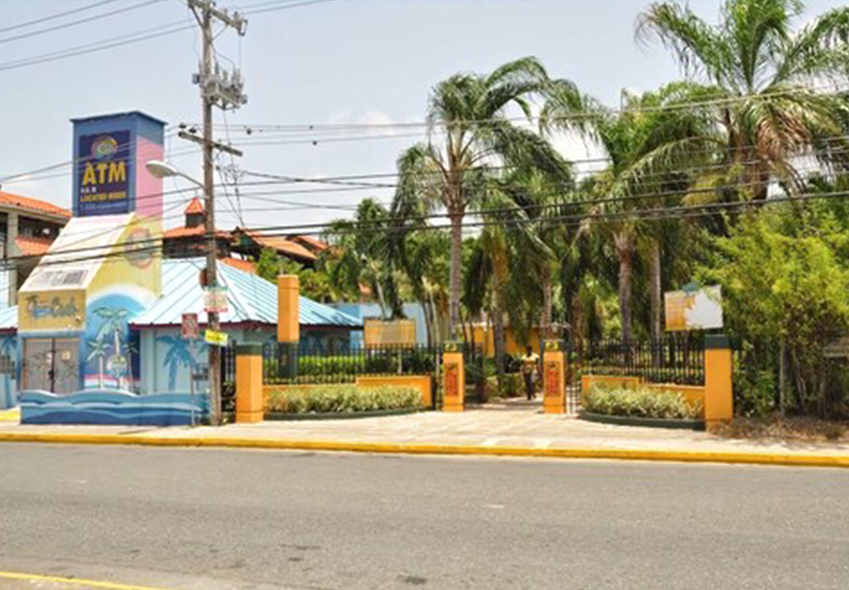 A gated entrance with palm trees, a blue ATM sign, and a building with a colorful mural.