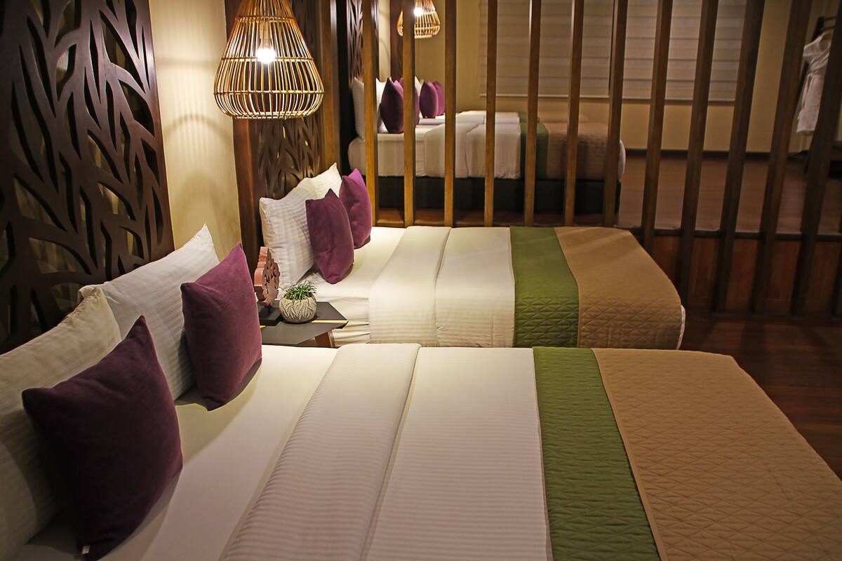Habitación de hotel con dos camas, cada una con una colcha verde y blanca, almohadas moradas y una pequeña mesa con una planta. Hay lámparas de mimbre sobre las camas y un panel de madera al fondo.