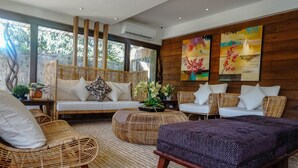 A modern living room with wicker furniture, a sofa, and a coffee table. There are two paintings on the wall and a large window with a view of greenery. - Balai Princesa (Puerto Princesa)