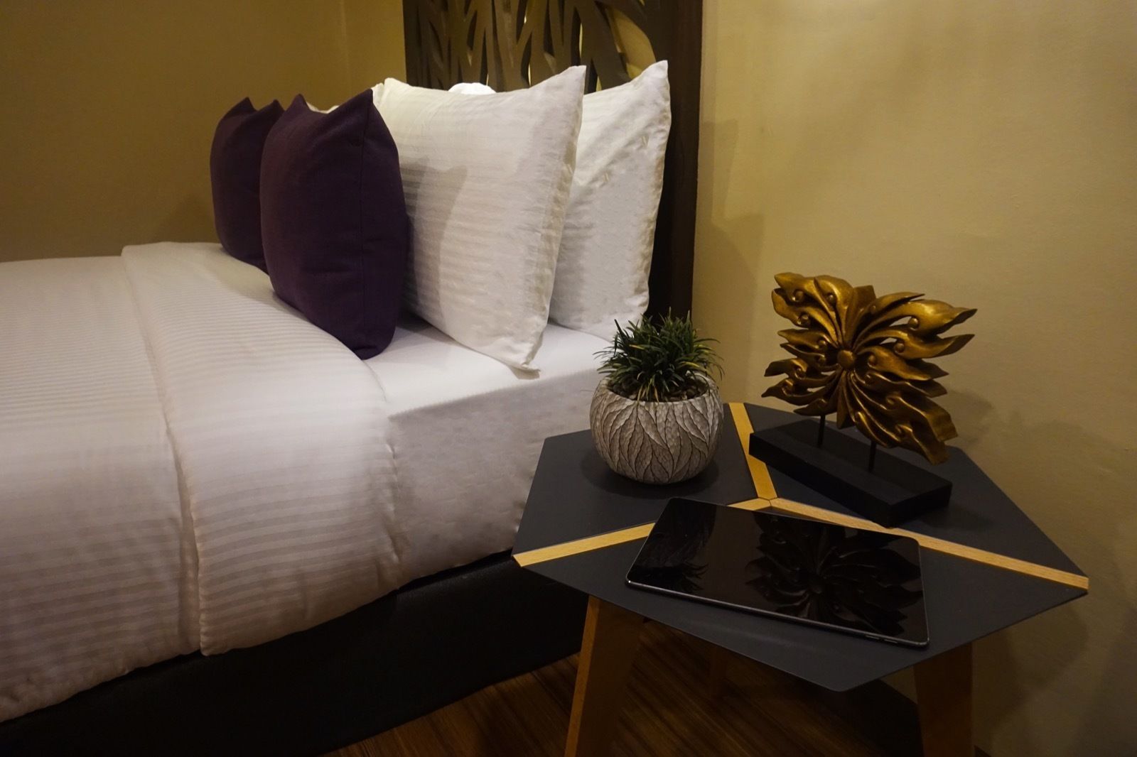 A neatly made bed with white bedding and purple pillows, a small potted plant, and a decorative golden sculpture on a black side table.