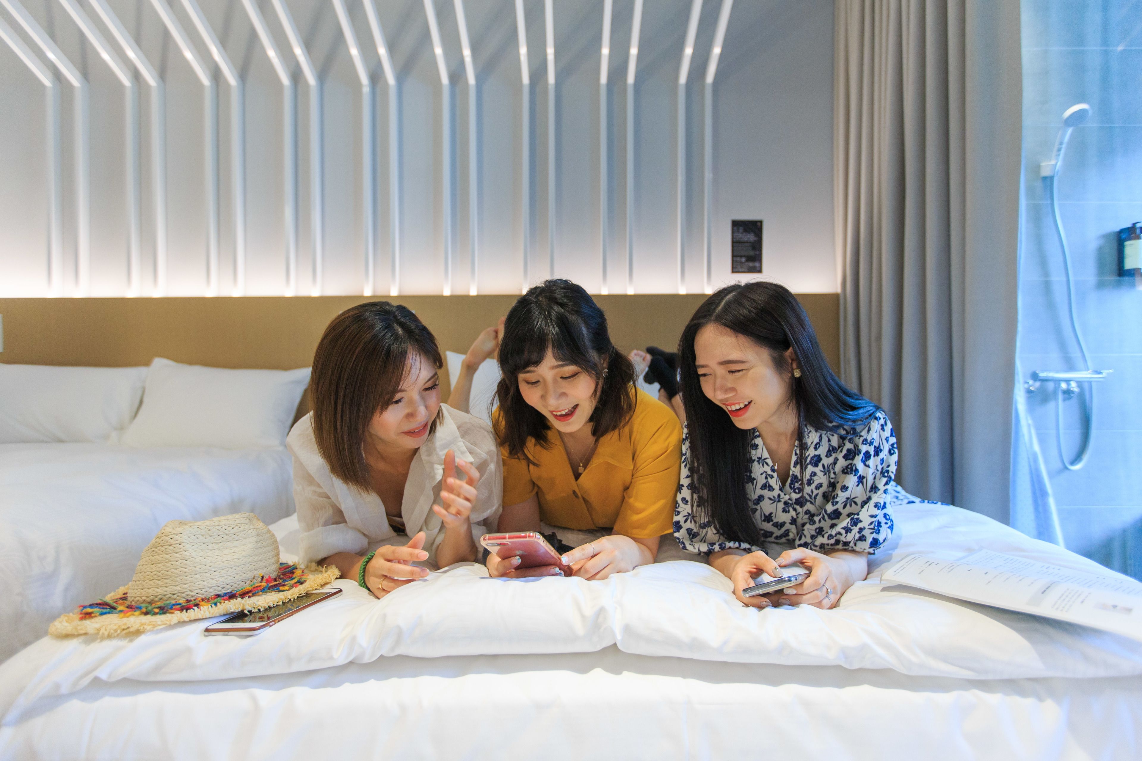 Three people lying on a bed in a hotel room with a modern design, a straw hat, and a smartphone on the bed.
