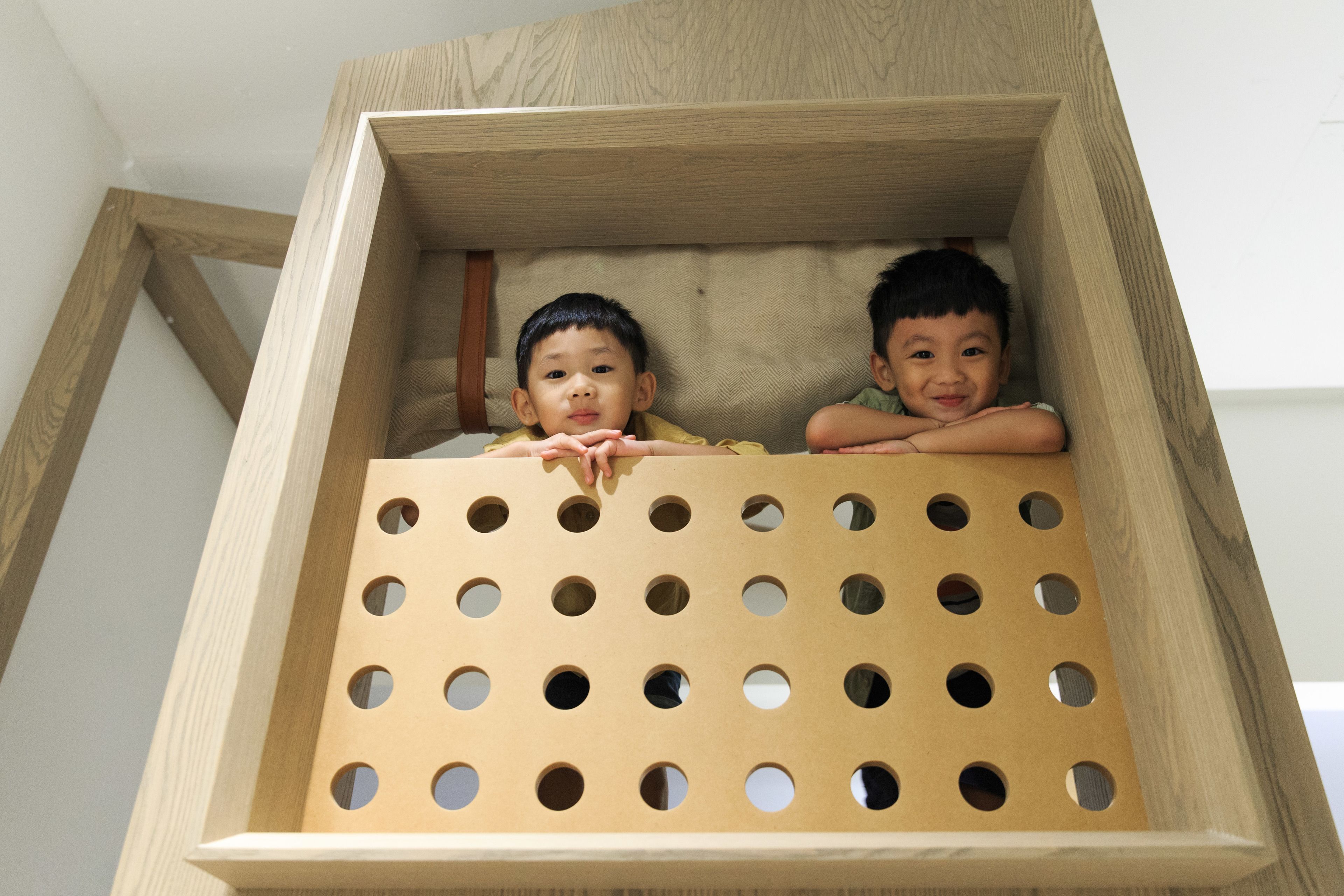 Two children in a wooden playhouse with a grid of holes on the front.