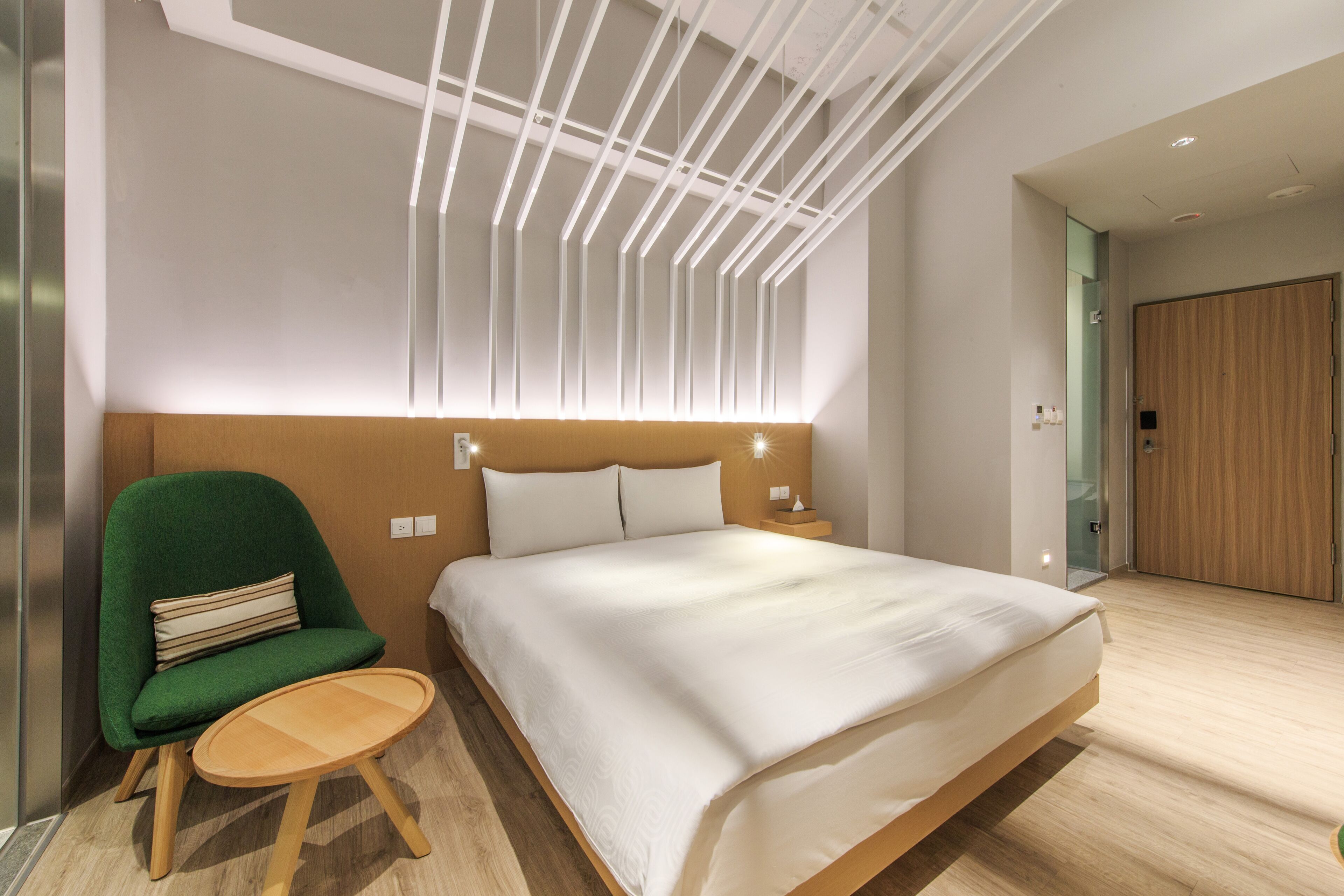 Una habitación de hotel moderna con una cama grande, un sillón verde, una pequeña mesa de madera y una lámpara de techo original.