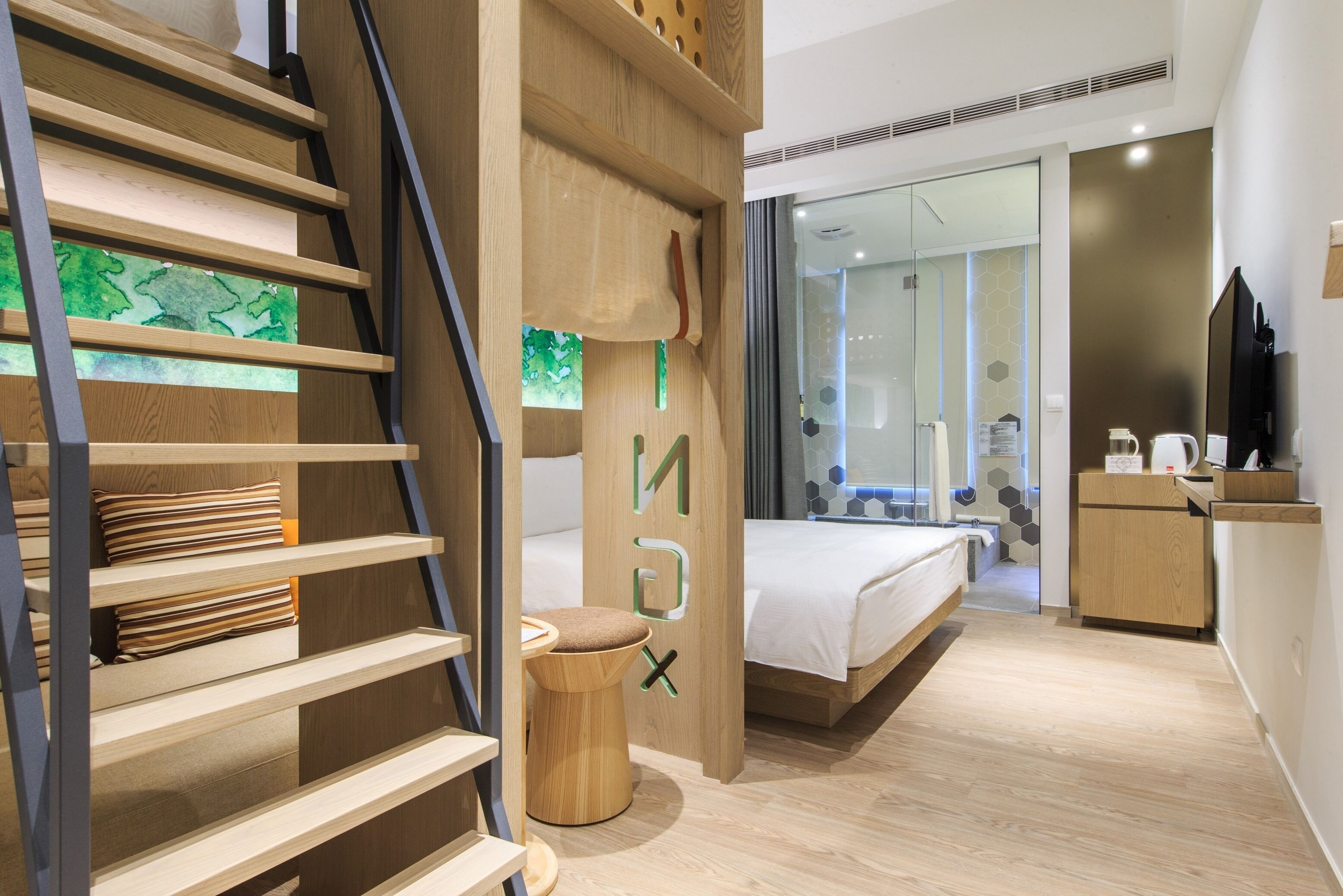 Une chambre d’hôtel moderne avec un escalier en bois, un lit doté d’un matelas blanc, un petit tabouret en bois et une porte en verre donnant sur une salle de bain.