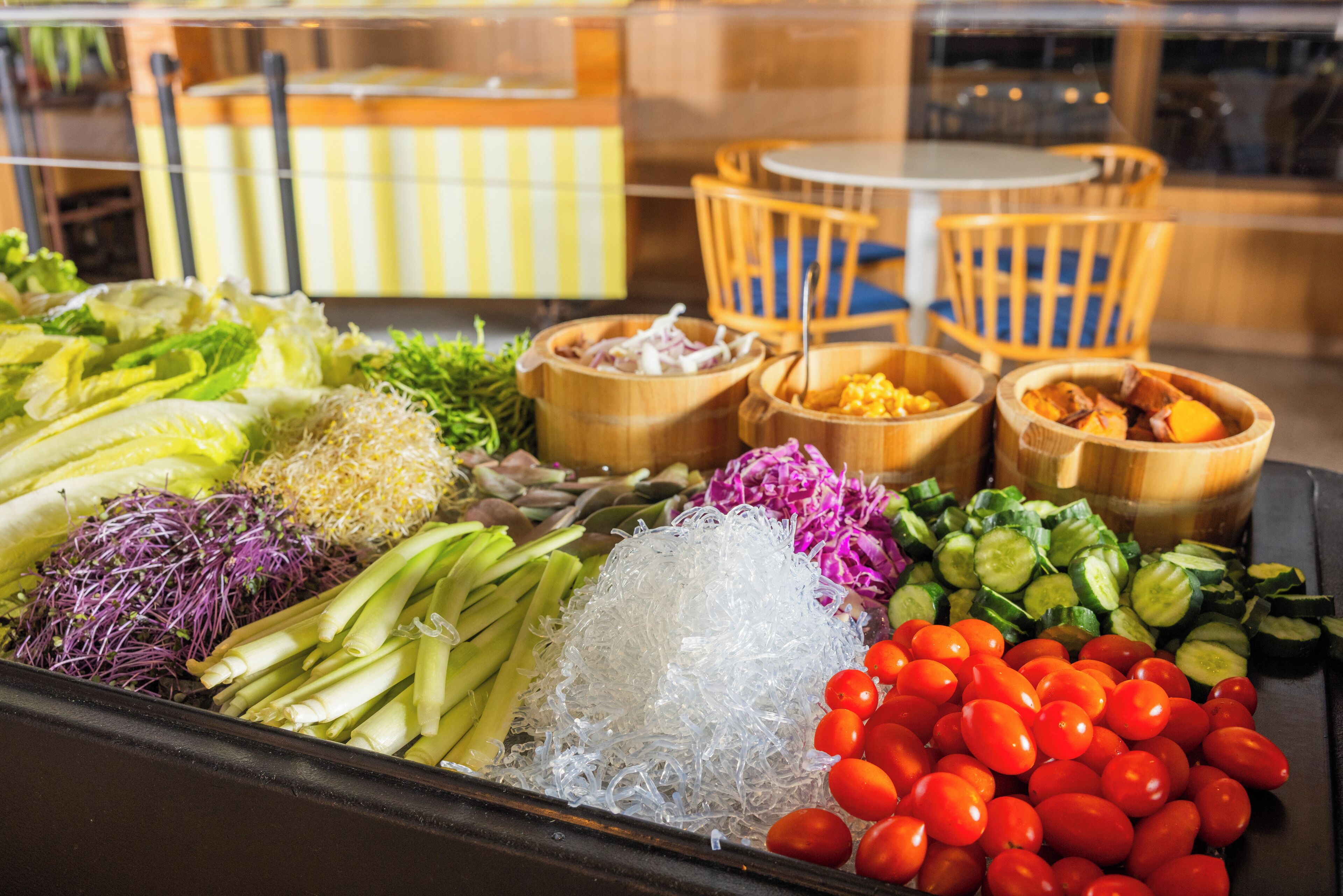 A variety of fresh vegetables and herbs displayed in wooden bowls, with a dining area in the background.