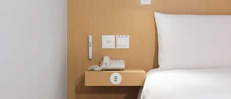 A hotel room with a wooden headboard, a bedside table with a phone, a power outlet, and a thermostat.