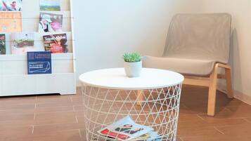 Una mesa redonda y blanca, con estructura de alambre, que sostiene una planta en maceta, una silla con cojín color beige y una estantería con varios libros y revistas.
