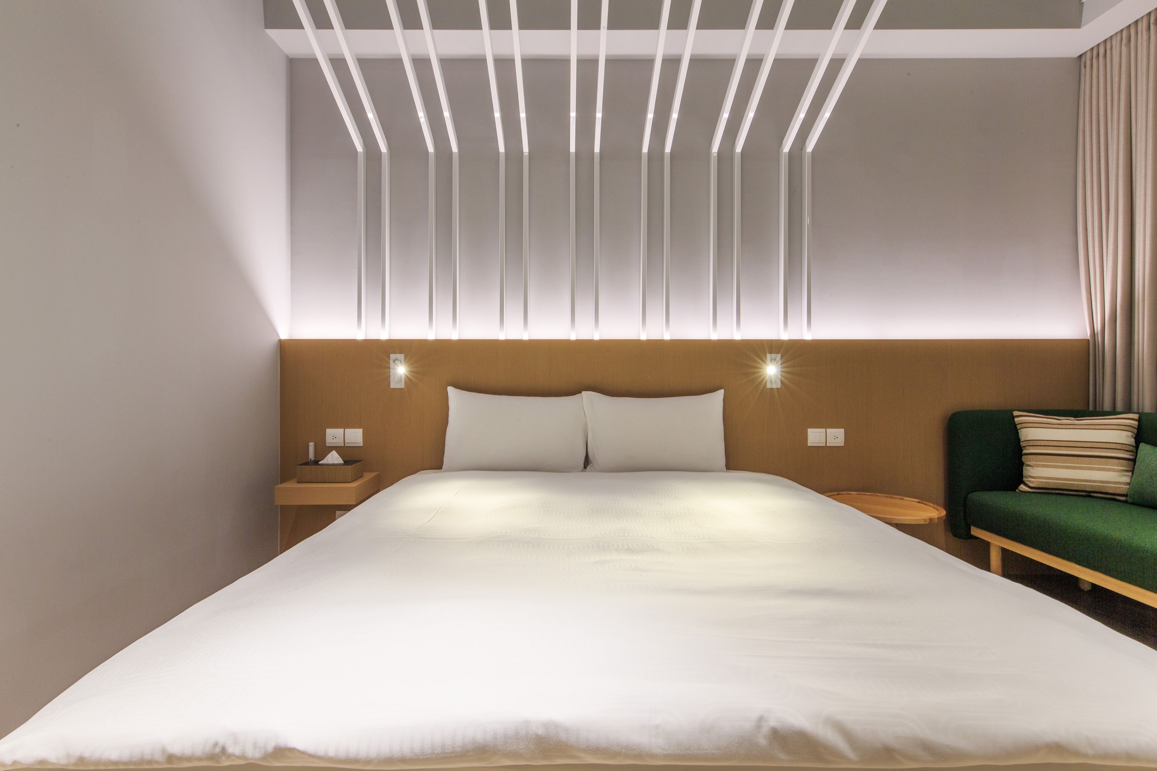A modern hotel room with a large bed, a wooden headboard, bedside tables, and a green sofa.