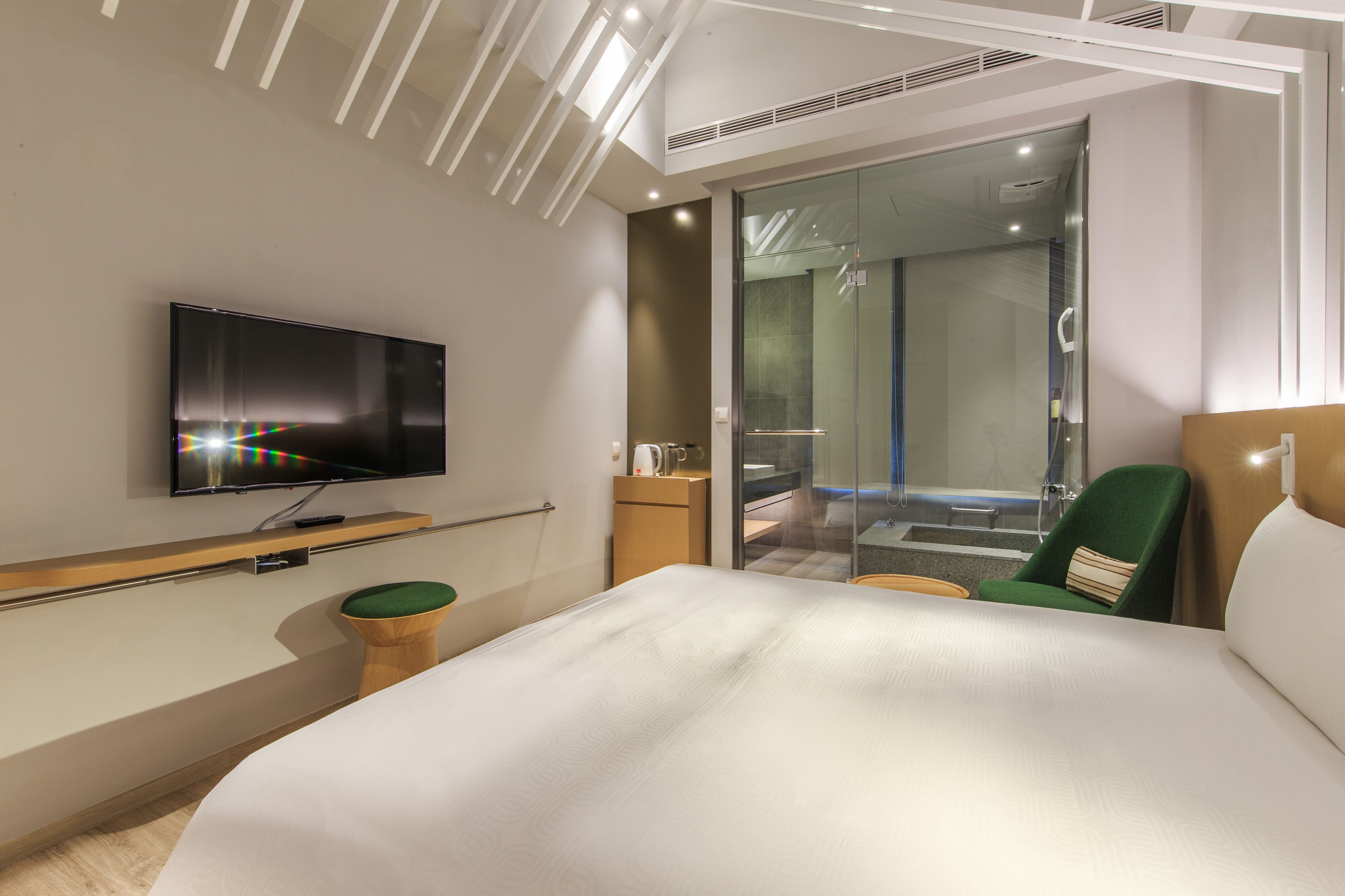 Habitación de hotel moderna con una cama grande, un televisor de pantalla plana, un taburete de madera, una silla verde y una zona de ducha con mampara de cristal.