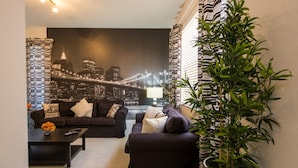 Un salon moderne avec vue sur la ville, deux canapés sombres, une table basse et une plante en pot.