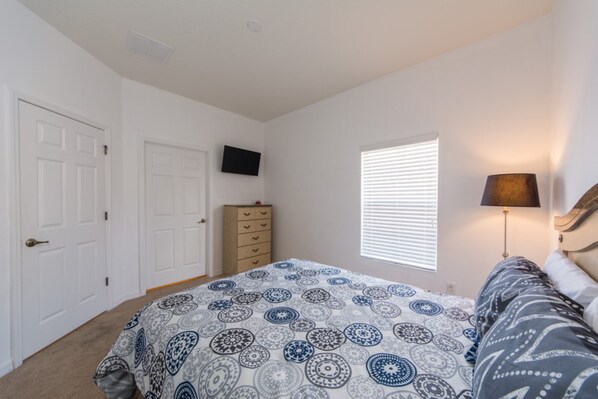 Un dormitorio con cama, cómoda, televisor en la pared y ventana con persianas.