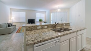 A modern kitchen with granite countertops, a double sink, and white cabinetry. There is a dining area with a sofa and a TV in the background.