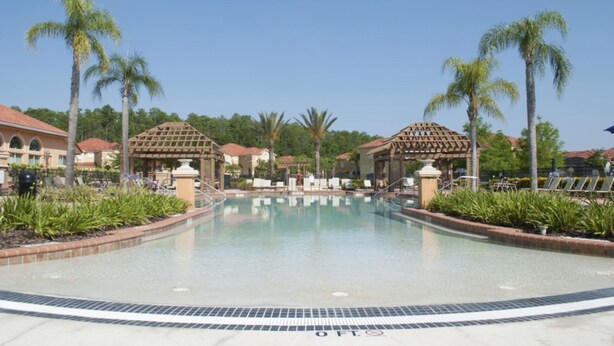 A pool area with lounge chairs and palm trees, surrounded by buildings with red-tiled roofs.