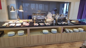 A buffet setup with various food items and utensils, a person in the background, and a view of the outside through a window.