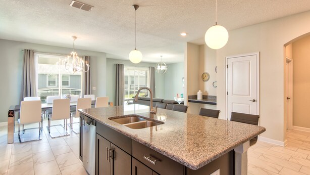 A modern kitchen with granite countertops, stainless steel appliances, and a large island. There are bar stools and a dining table in the adjacent dining area. - Champions Gate Home 2 U- Thor - Cg1561 (Davenport)