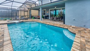 Una piscina con solárium de baldosas, un porche cubierto con mobiliario y una vista de una casa al fondo.