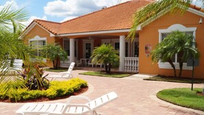 Un bâtiment doté d’un toit en tuiles et d’un porche couvert avec des colonnes blanches. On y trouve des espaces de détente en plein air, avec des chaises et des tables blanches.