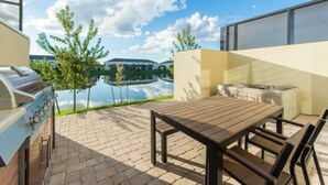 An outdoor patio with a grill, table, and chairs, overlooking a body of water and buildings.