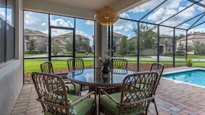 A covered patio with a glass table surrounded by wicker chairs, overlooking a swimming pool and residential buildings. - Champions Gate Home 2 U- Zeus (Davenport)