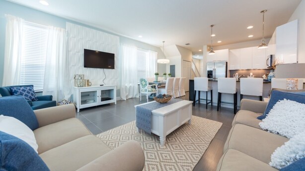 A modern living room with a sofa set, a coffee table, and a TV mounted on the wall. There is a kitchen area with bar stools and a dining table. - Serenity Home 2 U- Winter (Clermont)