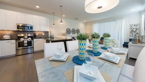 A modern kitchen with stainless steel appliances, a dining area with a table set for four, and a living area with a sofa and decorative plants. - Serenity Home 2 U- Winter (Clermont)