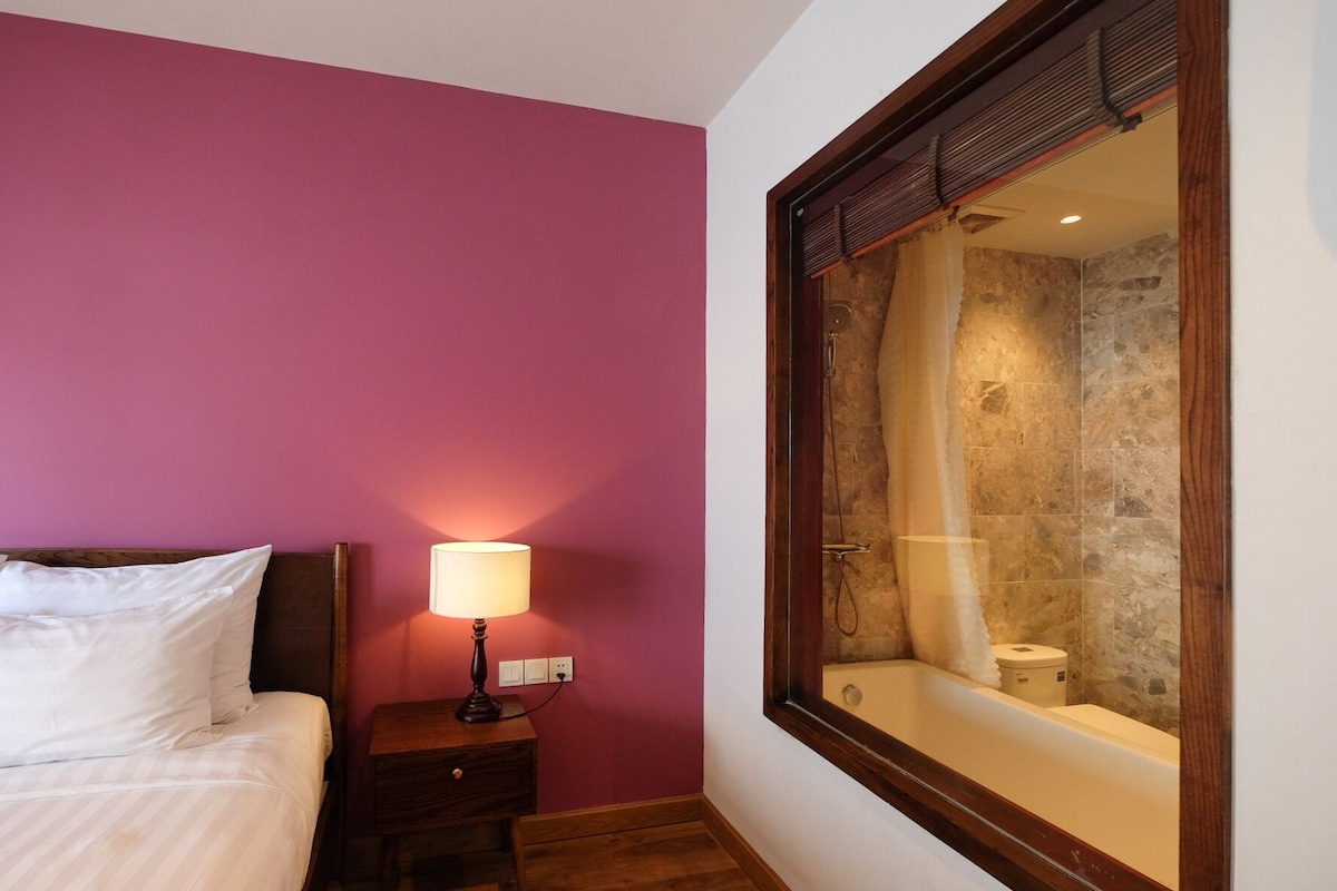 Hotelkamer met een bed, nachtkastje met lamp en een grote spiegel die een douchegedeelte weerspiegelt.