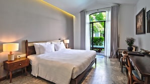 A hotel room with a large bed, bedside lamps, a desk with chairs, and a view of greenery outside.