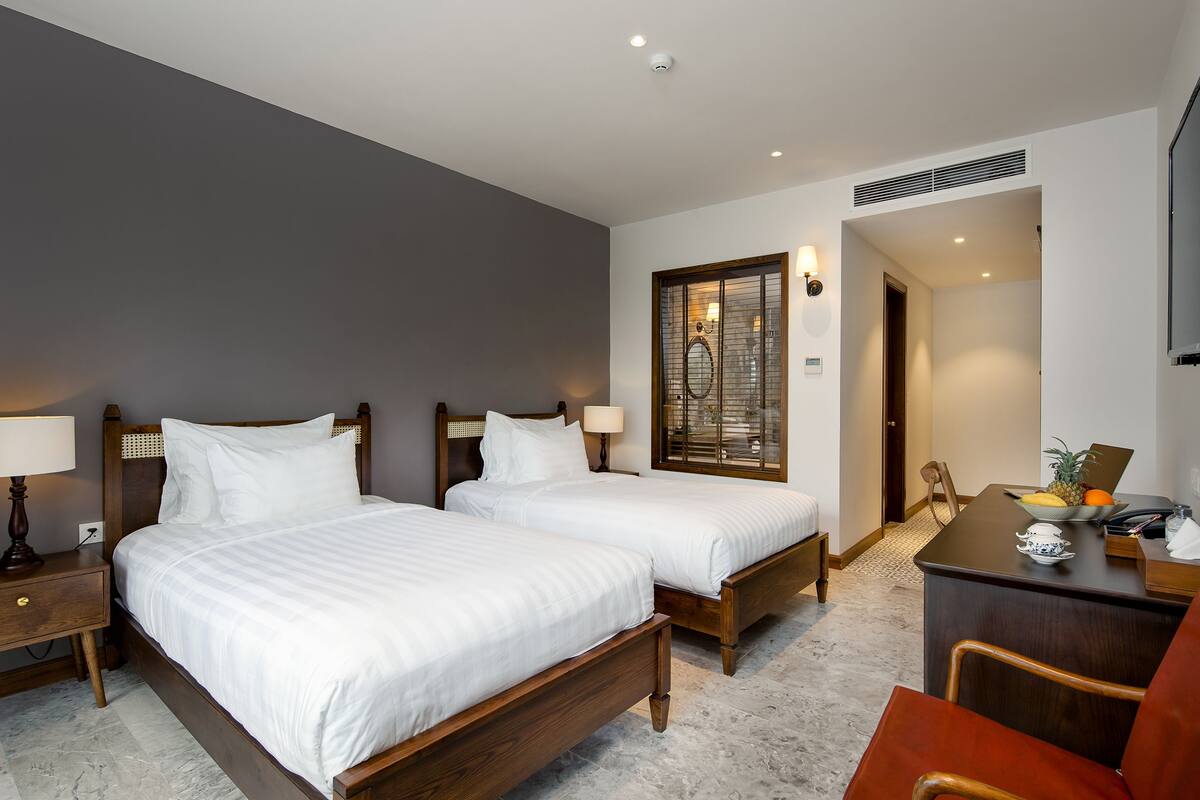 Een hotelkamer met twee houten bedden, een bureau met een laptop, een stoel en een klein tafeltje met fruit.