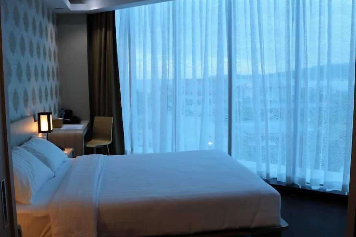 Habitación de hotel con una cama grande, un escritorio con lámpara, una silla y vistas de la ciudad a través de cortinas translúcidas.