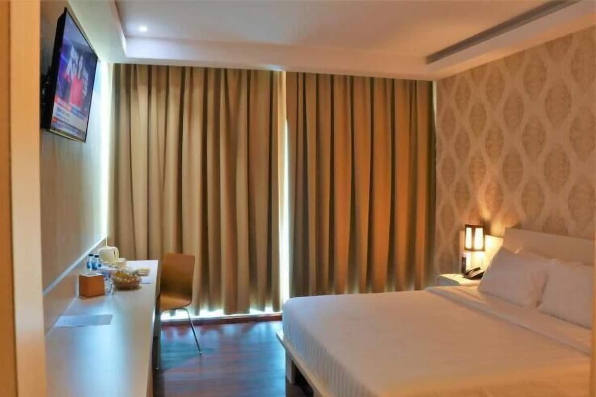 Habitación de hotel con cama, escritorio con silla, televisor montado en la pared y ventana con cortinas.