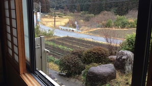 A view from a window overlooking a road with a bench, a fence, and a hillside with trees. - Minshuku Ryokan Kawai (Shinshiro)