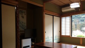 A traditional Japanese room with tatami flooring, a low table, a television, sliding doors, and a view of a forest through a window. - Minshuku Ryokan Kawai (Shinshiro)
