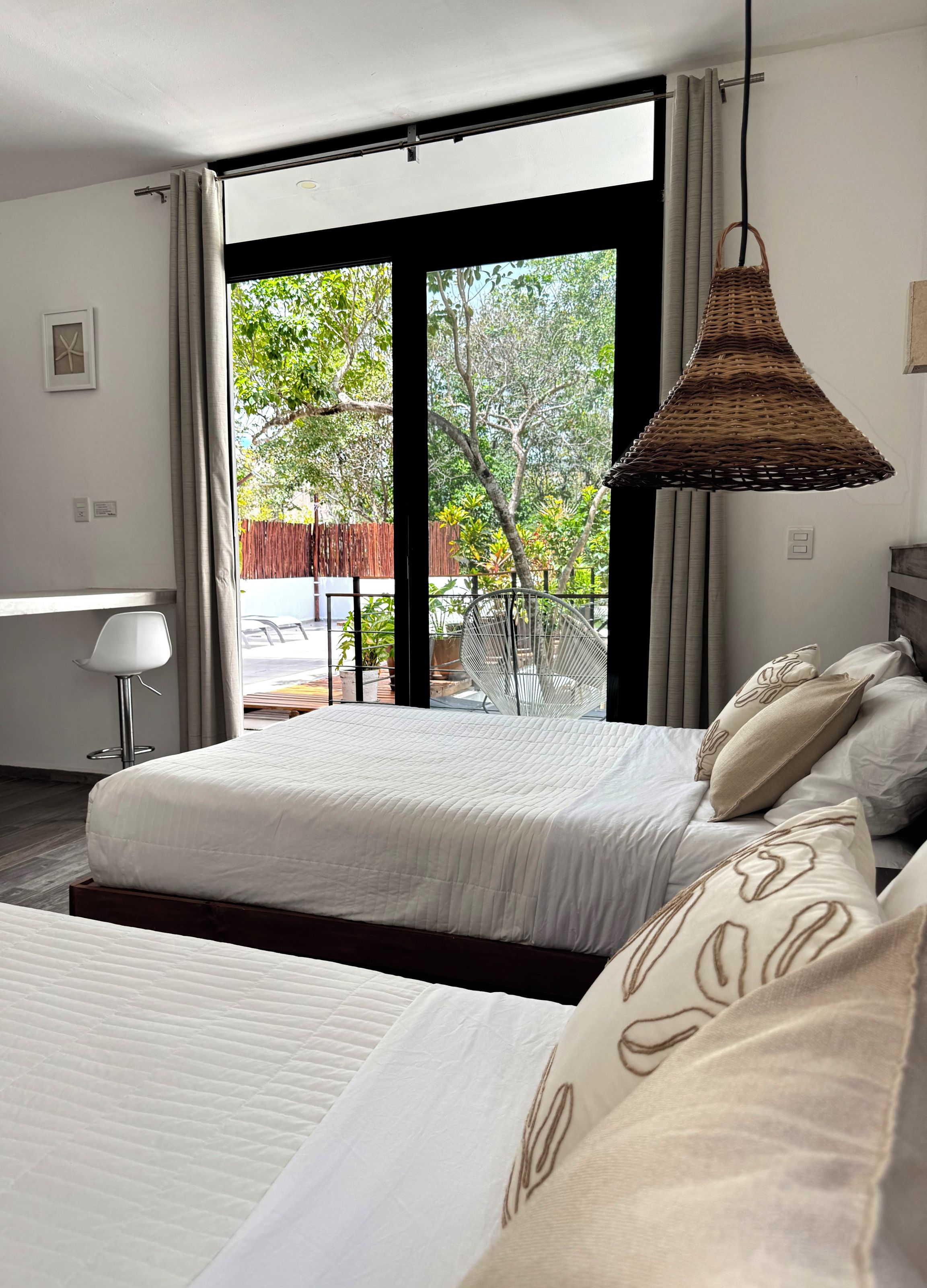 A hotel room with two beds, a large window with a view of greenery, and a hanging wicker lamp.