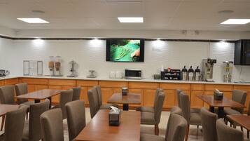 Un café moderno con mesas y sillas de madera, un televisor de pantalla plana montado en la pared, y varias máquinas de café y electrodomésticos en la barra.