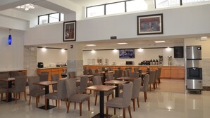 A spacious dining area with multiple tables and chairs, a kitchen area with a refrigerator, and a large window providing natural light. - Lic Plaza Hotel (Long Island City)