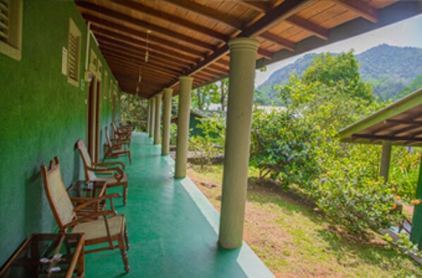 A covered corridor with wooden chairs and pillars, leading to a lush green landscape with mountains in the background. - Blue Magpie Lodge (Kalawana)
