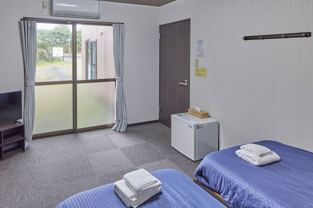 A hotel room with a television, two beds with blue bedding, a small refrigerator, and a sliding glass door leading to an outdoor area.