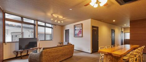 A modern hotel room with a large wooden dining table, wicker sofa, and a flat-screen TV.