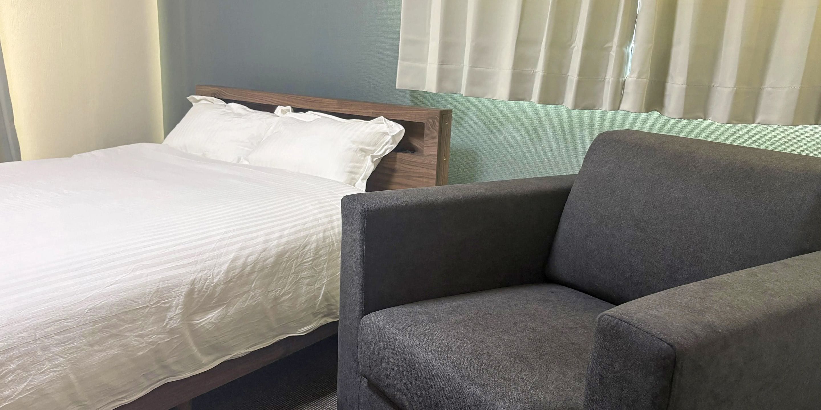 A bed with white bedding and a grey upholstered armchair in a room with a window and blinds.