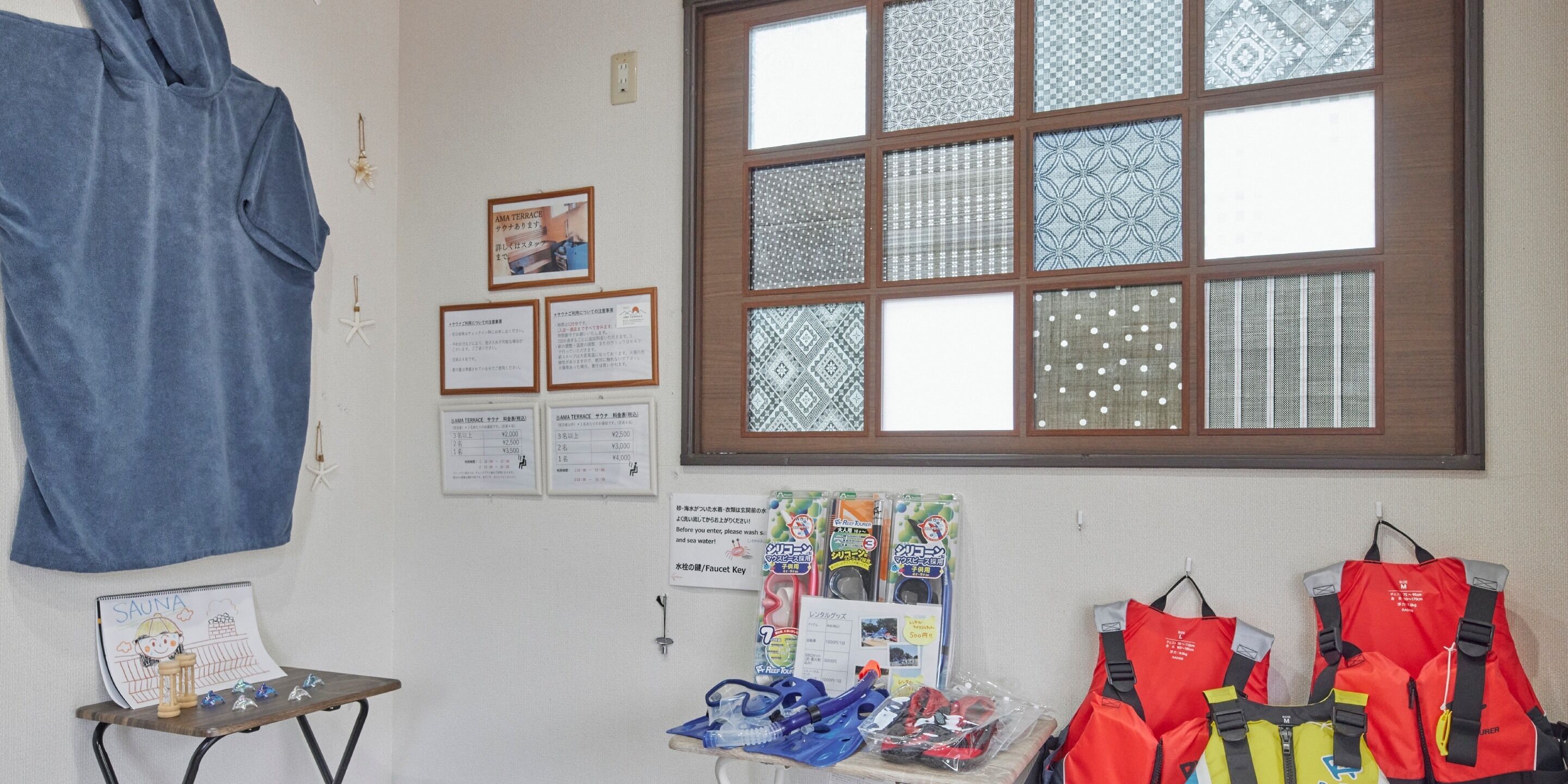 Una habitación con un ventanal grande, una toalla azul, una mesita con objetos y chalecos salvavidas rojos en la pared.
