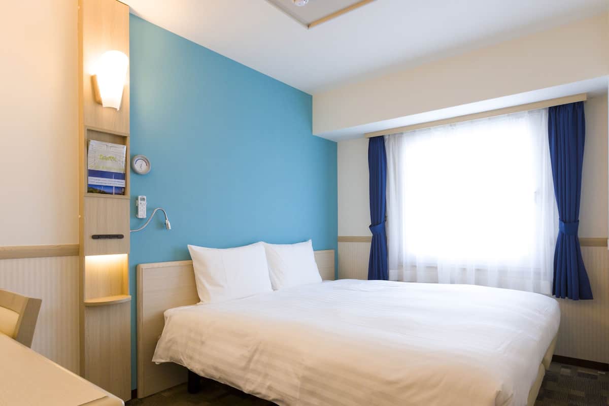 A hotel room with a bed, a desk, a wall-mounted phone, and a window with curtains.