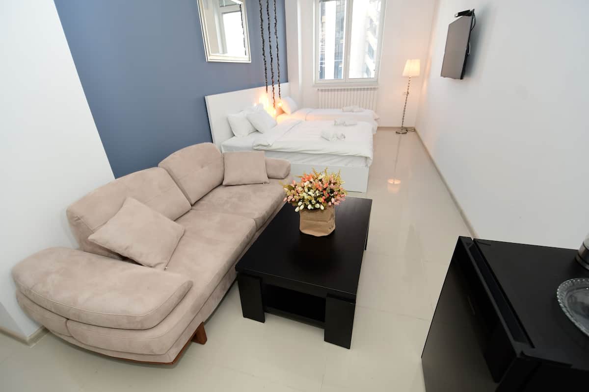 Ein modernes Wohnzimmer mit einem beigefarbenen Sofa, einem schwarzen Couchtisch mit Blumen, einer schwarzen TV-Bank und einem Bett mit weißer Bettwäsche.