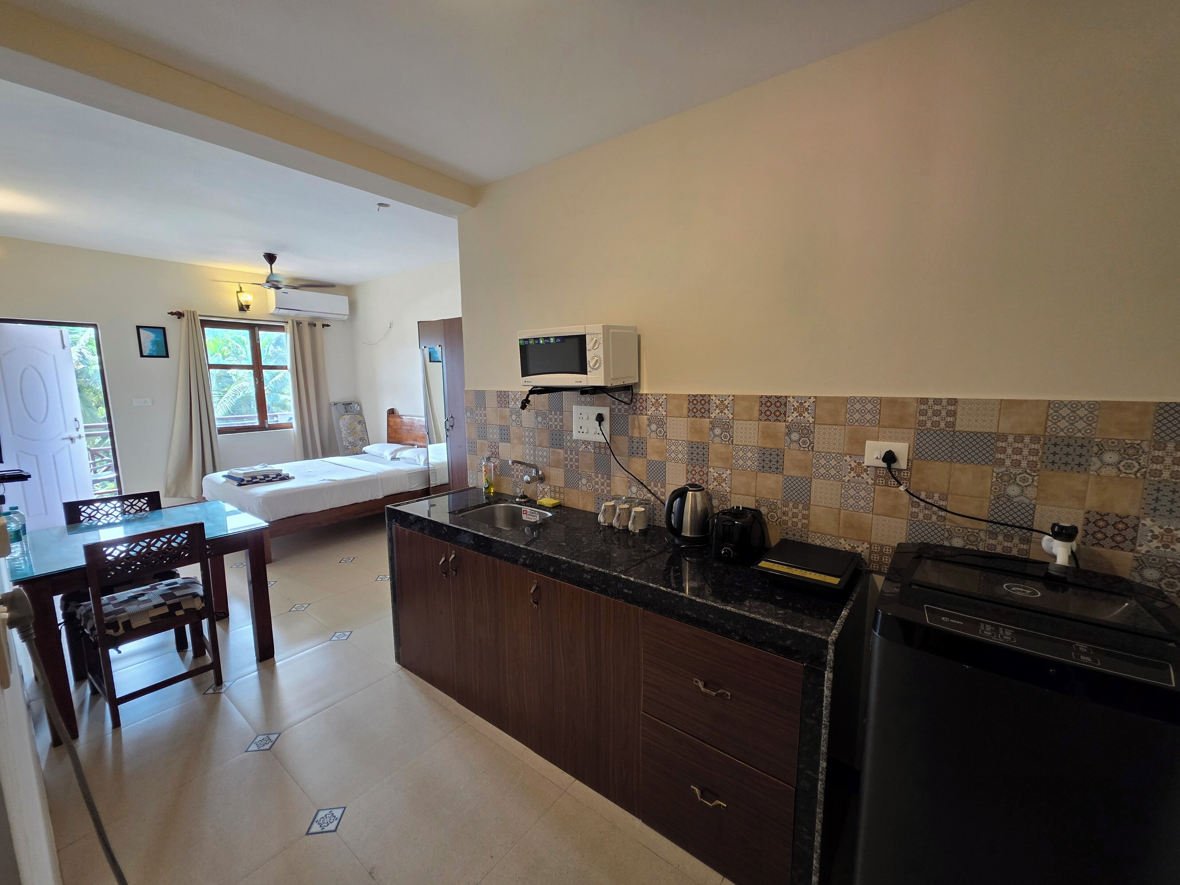 A compact kitchen with a microwave, sink, and a coffee maker. There is a bed in the adjacent room with a window and curtains.