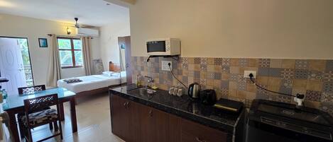 A compact kitchen with a microwave, sink, and a coffee maker. There is a bed in the adjacent room with a window and curtains.