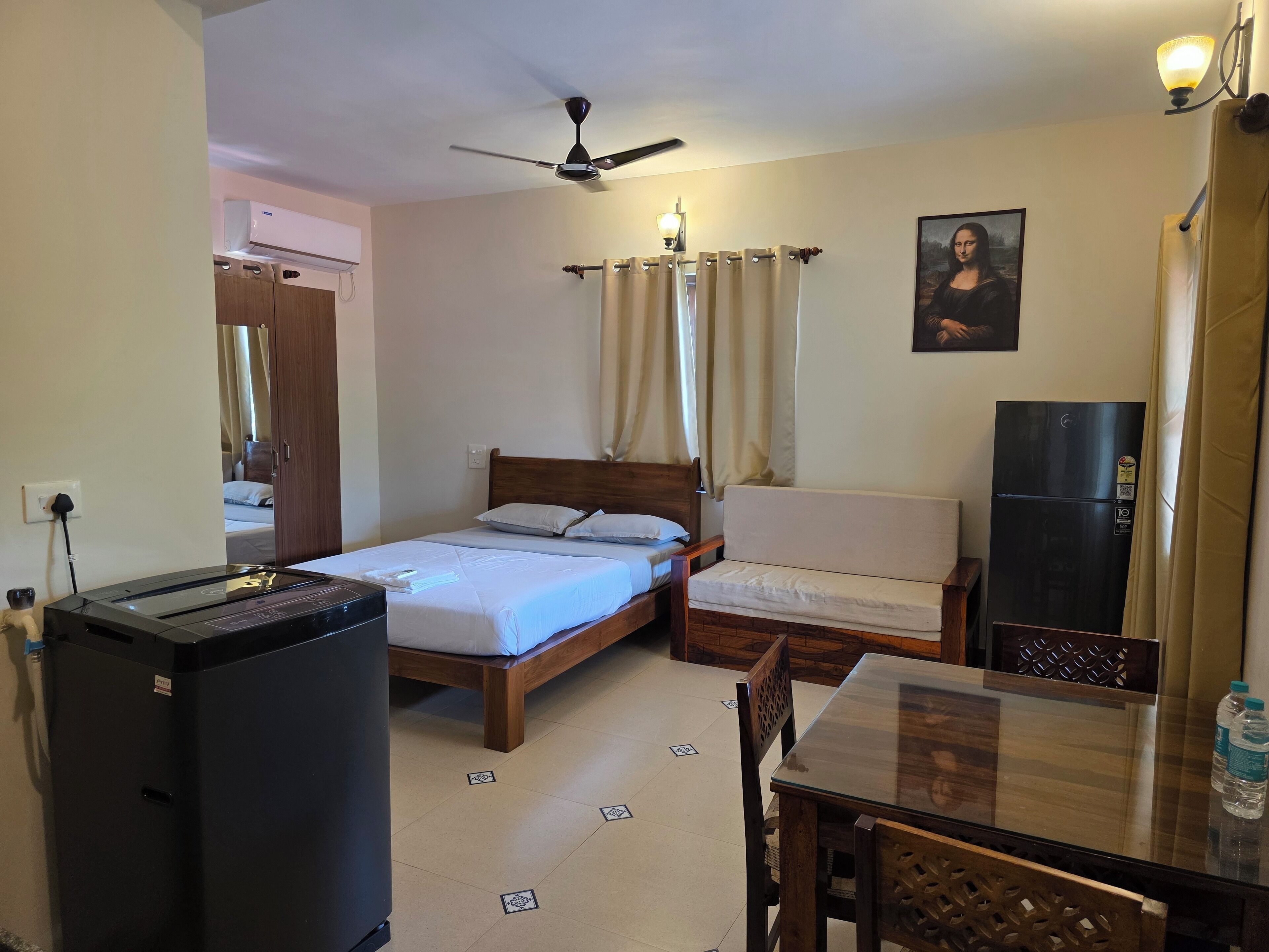 A hotel room with a bed, a chair, a table, a refrigerator, a ceiling fan, and a painting on the wall.