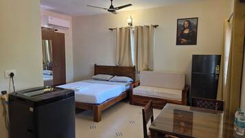 A hotel room with a bed, a chair, a table, a refrigerator, a ceiling fan, and a painting on the wall.