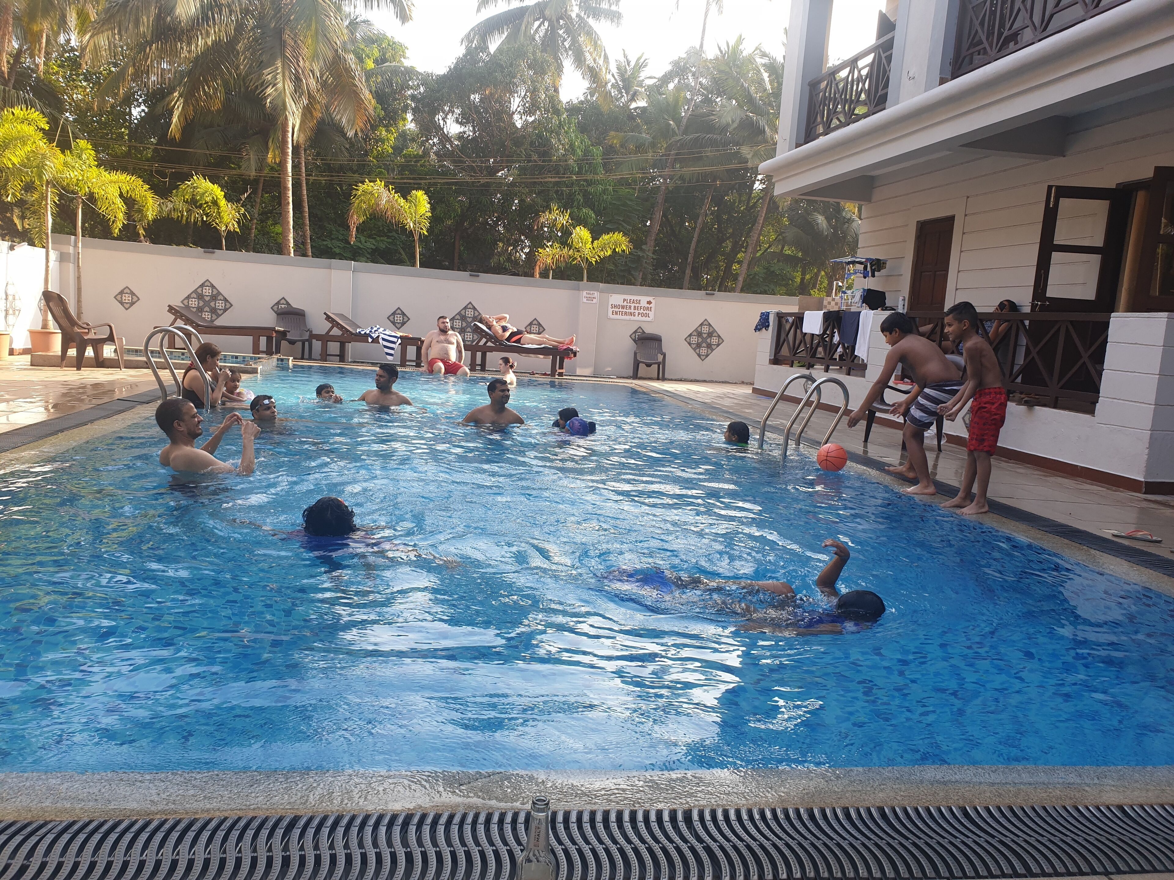 A swimming pool with people swimming and playing, surrounded by palm trees and a building with a sign that says "Please shower before entering pool."