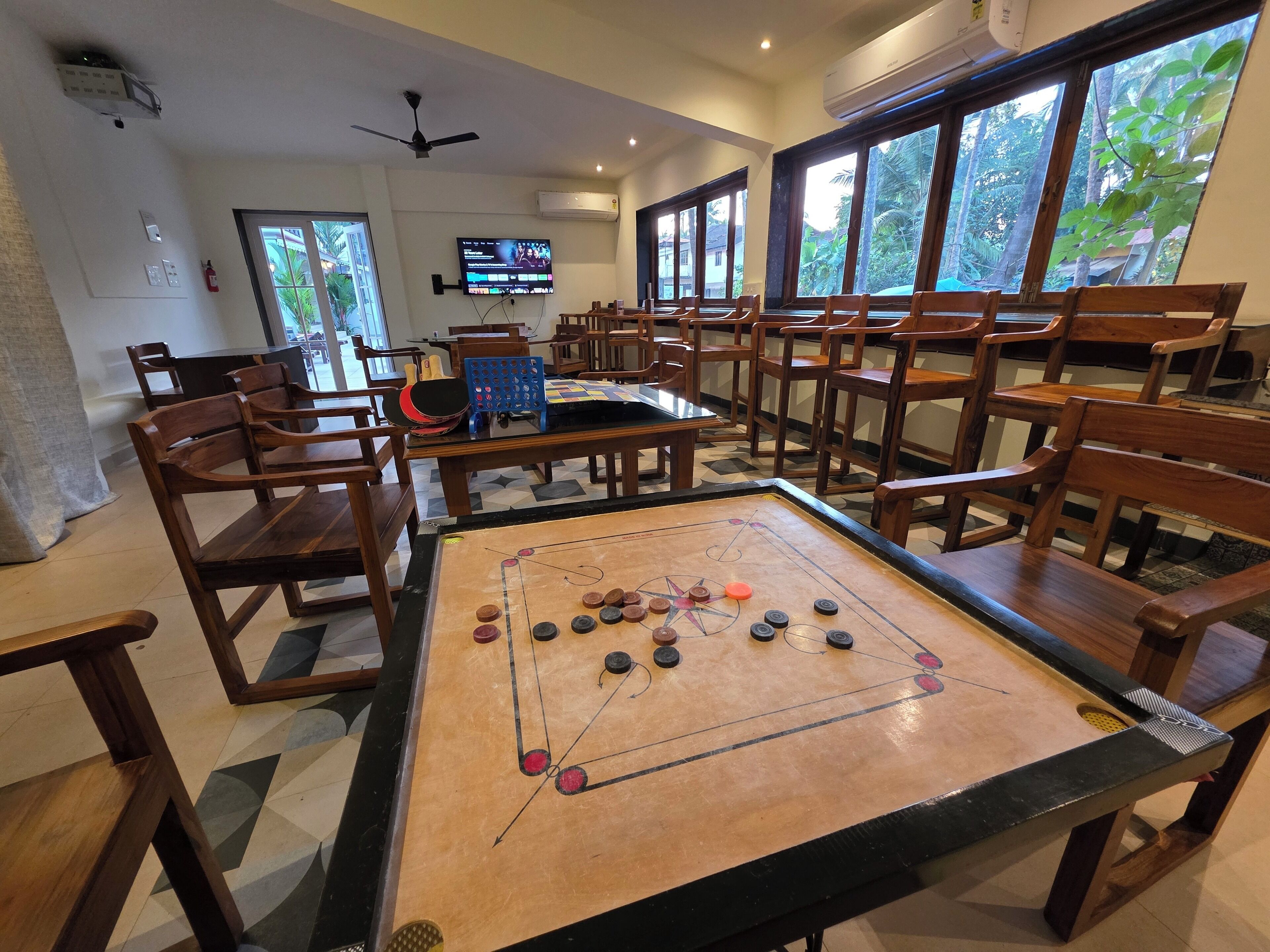 Una habitación con mesa de fútbol, sillas de madera, televisor y ventanas amplias con vista a follaje.