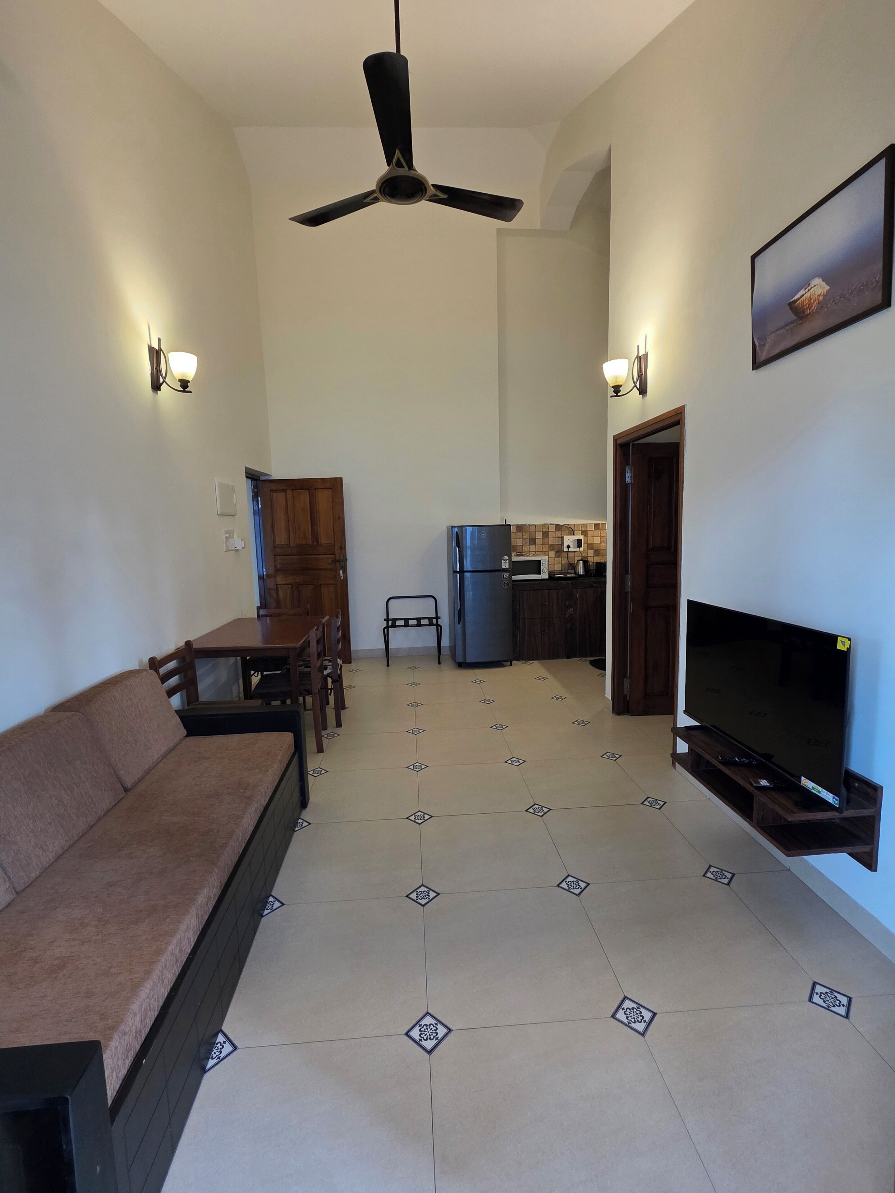 A living room with a sofa, TV, ceiling fan, and kitchen area with a refrigerator and microwave.